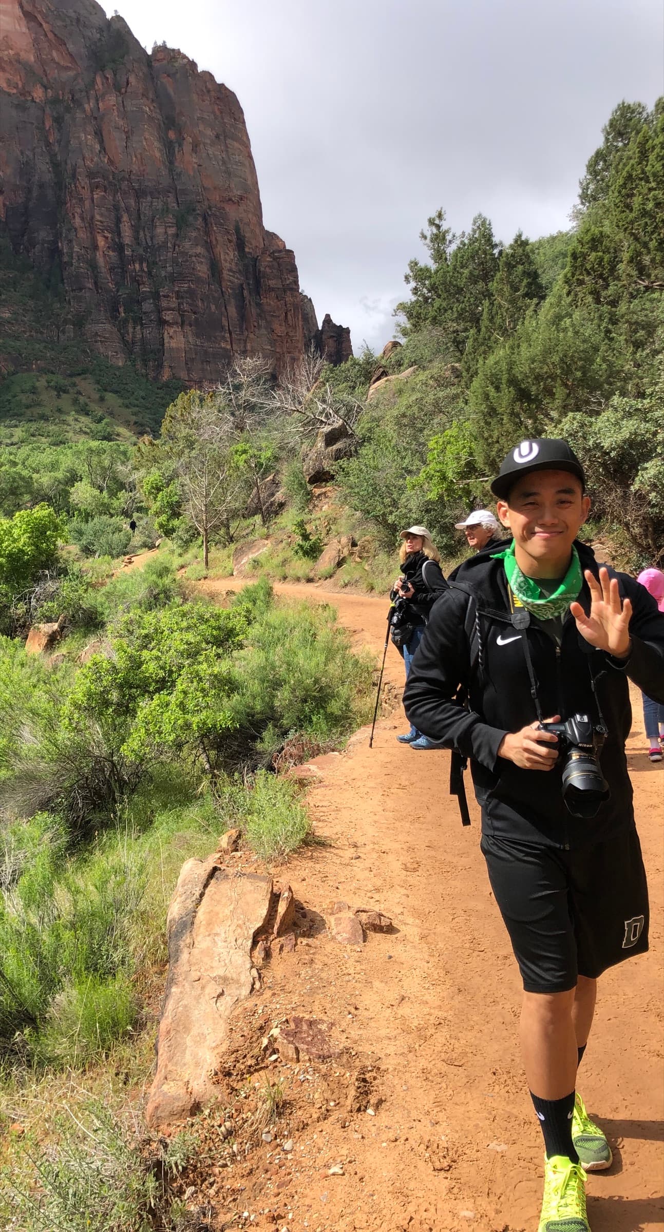 Valentino smiling and waving on a dirt hiking trail beneath the red rock cliffs of Zion National Park, a camera in his other hand.